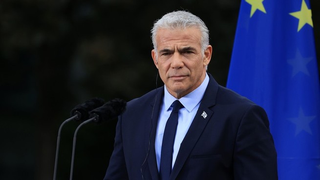 Yair Lapid, wearing a navy suit and tie, stands in front of a pair of microphones, with the EU flag in the background.