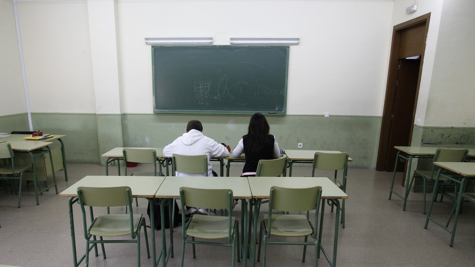 Two students sit at desks in an otherwise empty classroom in front of a chalk board.