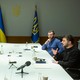 photo of large white meeting table with two women and a man on left and two men on right, one speaking, with Ukrainian flags in background