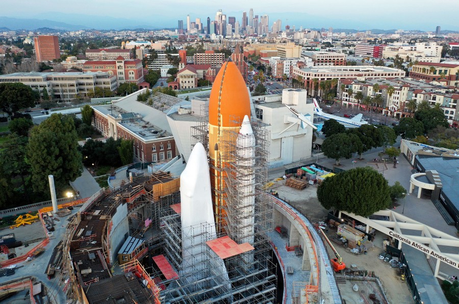 An aerial view of a space shuttle and booster rockets in a construction area, with the Los Angeles skyline in the background