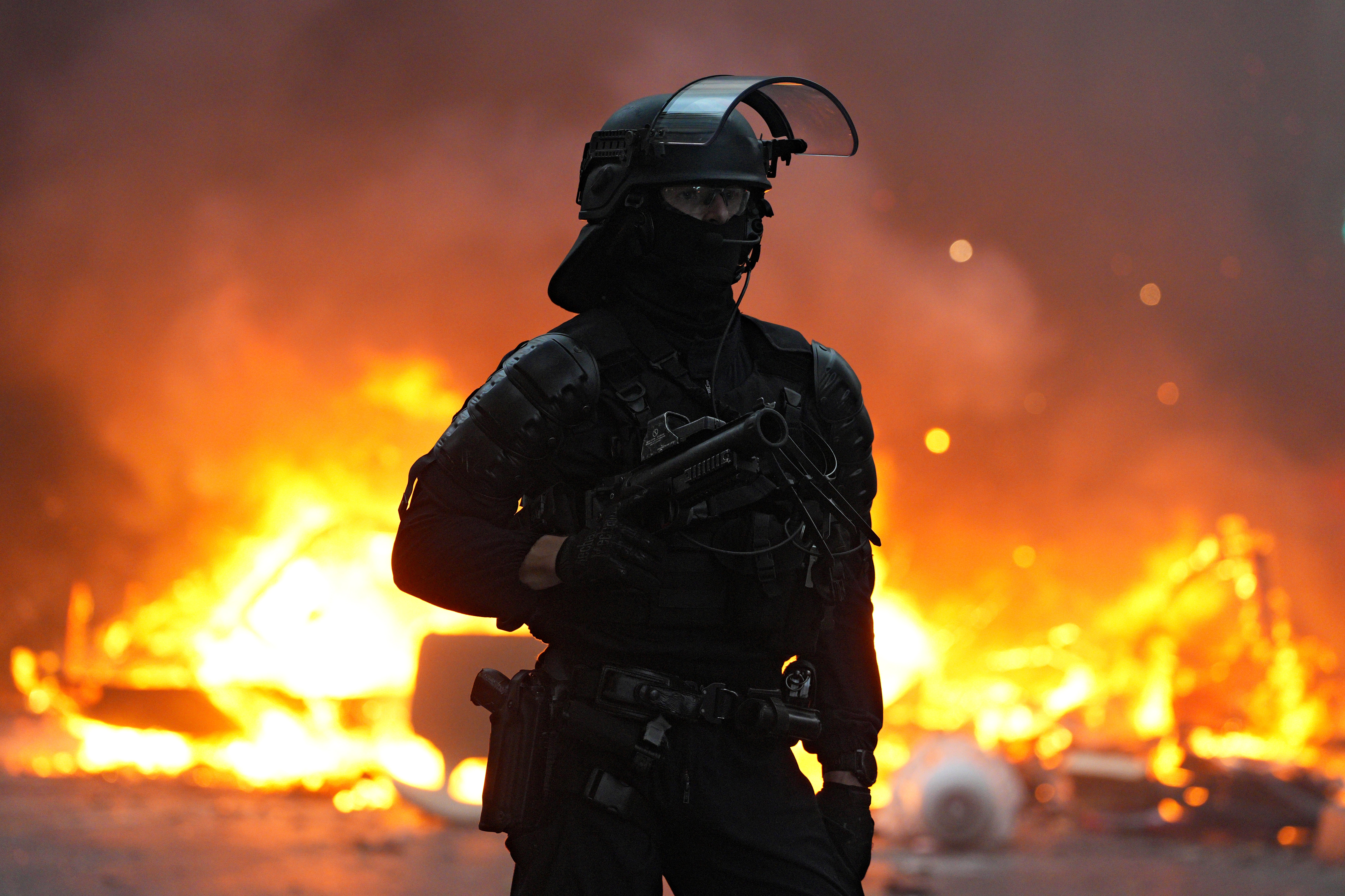 A police officer wearing riot gear stands in front of a burning barricade.