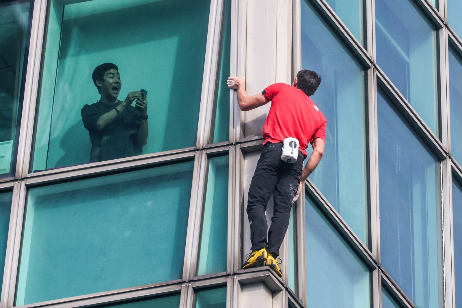 A person inside a tall building uses his phone to record a climber ascending the outside of the structure.