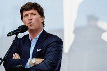 A photo of Tucker Carlson speaking at a microphone with his arms crossed