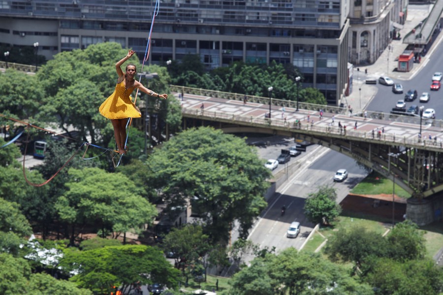 A tightrope walker balances on a wire high above a city boulevard.