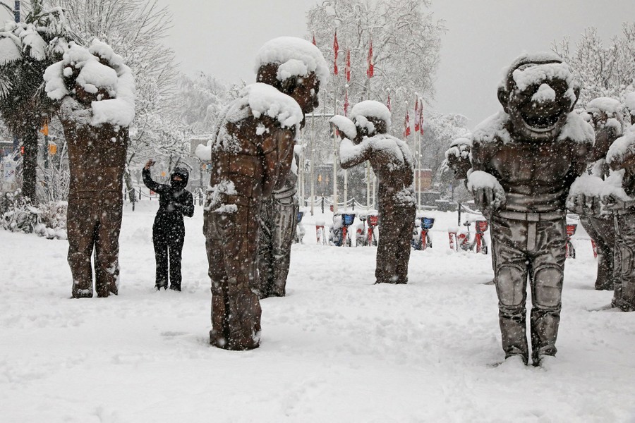 A person holds up a smartphone beside several snow-covered sculptures in a park.