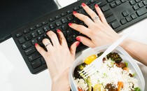 An overhead photograph of two hands typing on a keyboard, with a food container nearby on the desk
