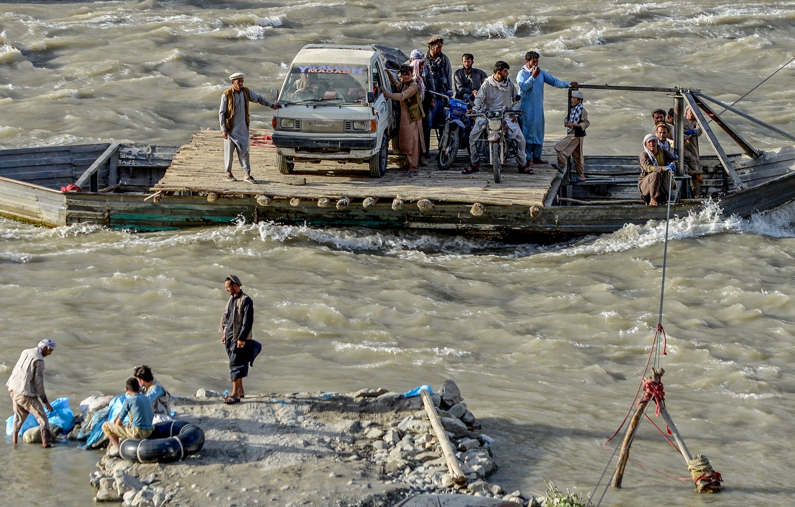 A group of people stand beside a small truck on a small wooden ferry , making their way across a swift-flowing river.