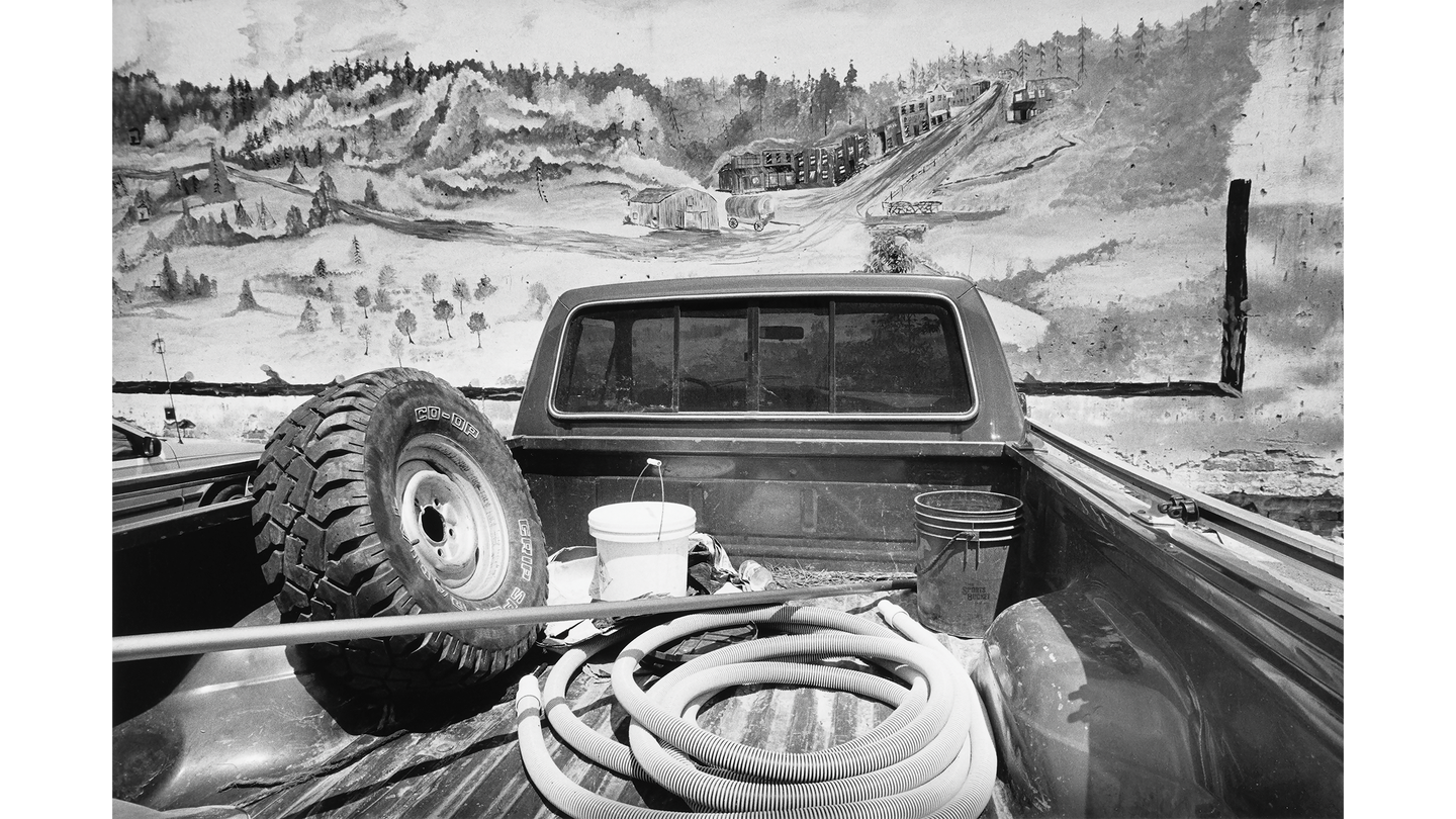 a photo of the rear of a pickup truck, its bed filled two buckets, a tire, and a hose, parked facing a mural of a mountain scene