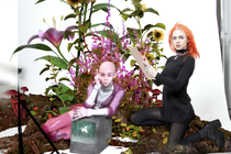 Grimes, in an orange wig, sits amid a field of flowers.