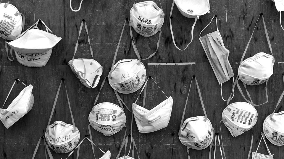 A black-and-white image of various N95 and surgical masks, hanging by their straps from pins on a wooden wall
