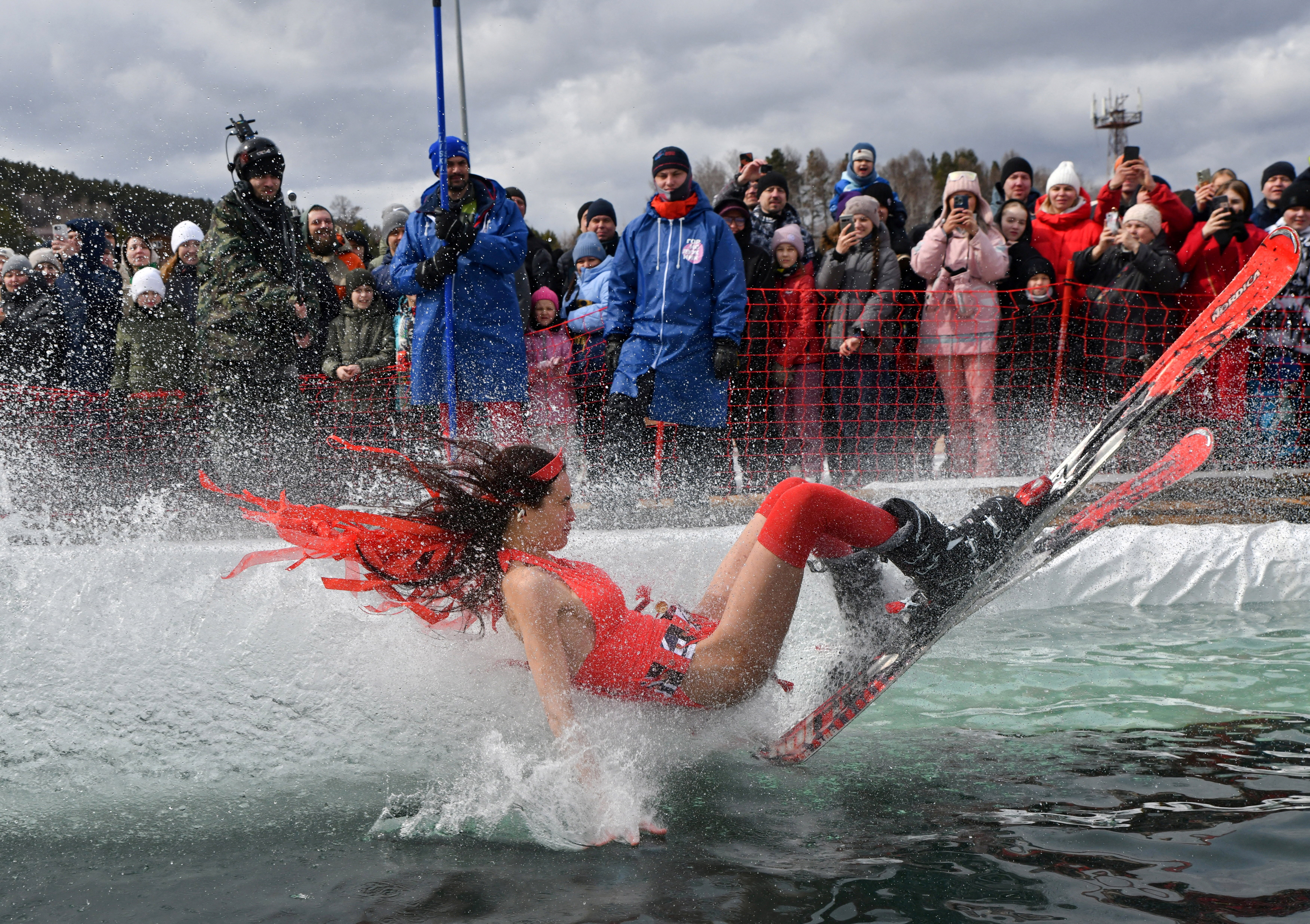 A skier in a festive costume leans back, splashing across a pond as a crowd watches.