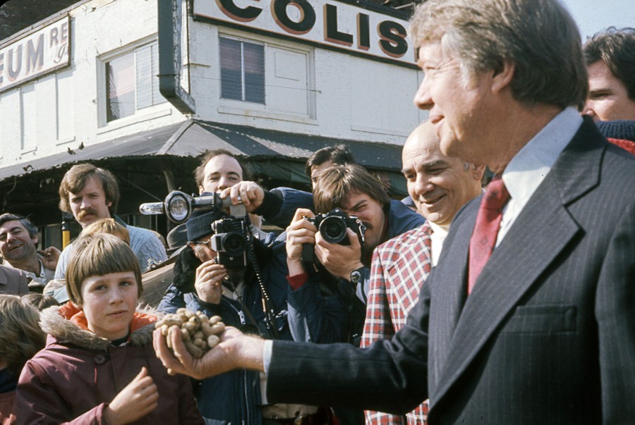 Jimmy Carter holds a handful of peanuts while speaking to a crowd of people and members of the press.