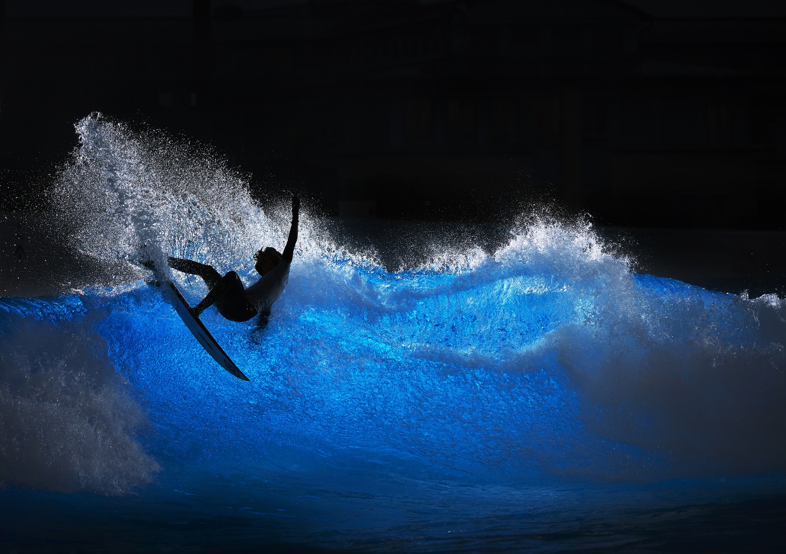 A surfer rides a wave at night, the blue water lit from behind in a wave pool.