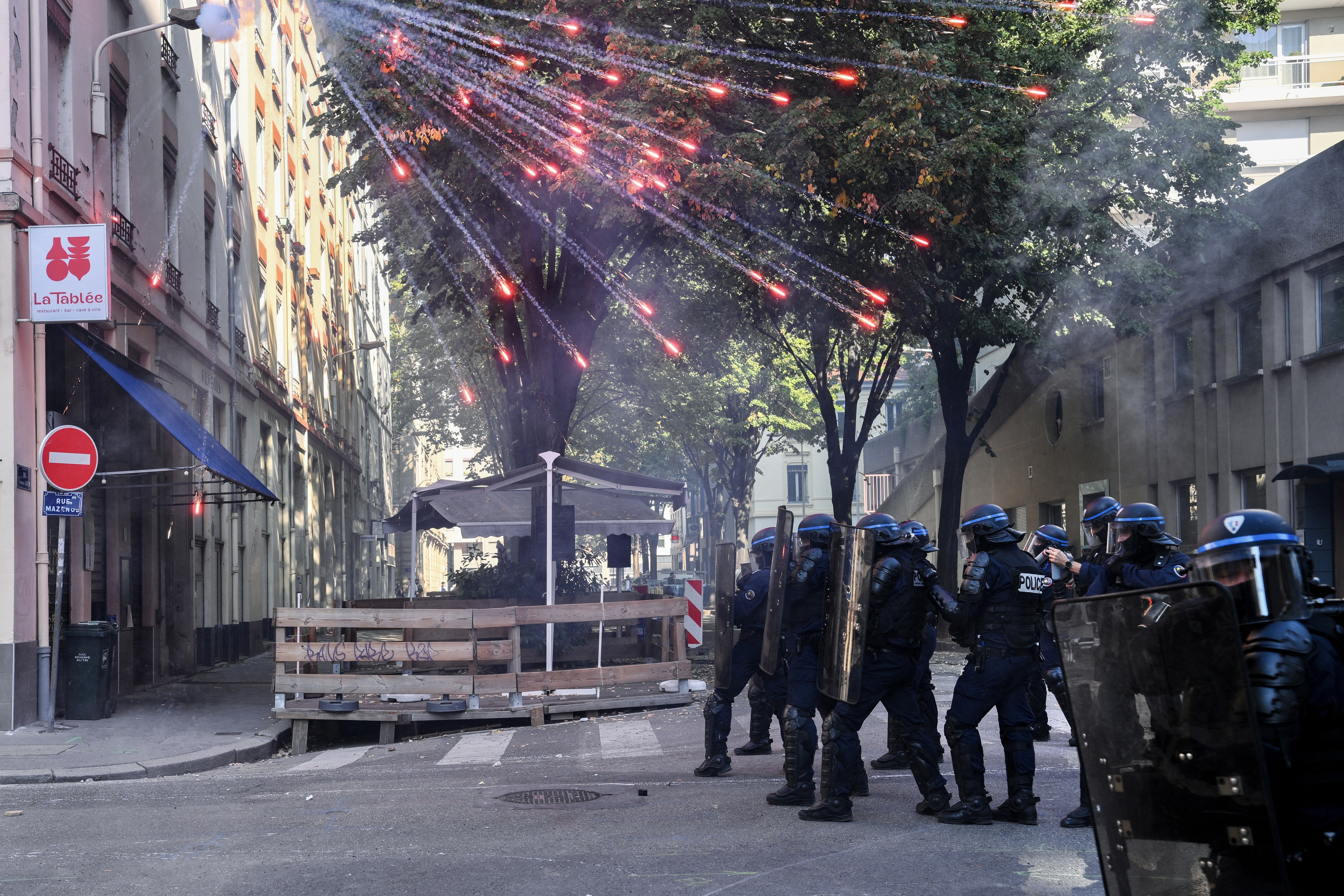 Riot police officers stand in a street as fireworks go off above them.