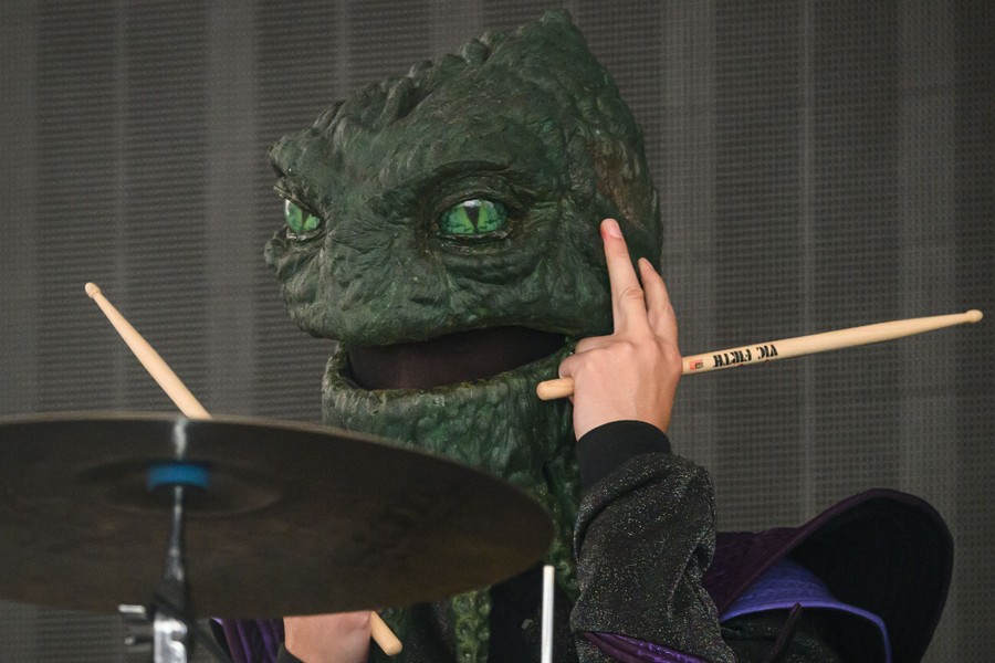 A close view of a drummer, onstage, wearing a full-head reptile mask