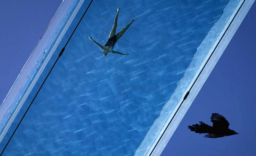 A woman swims as a bird flies underneath a transparent acrylic swimming-pool bridge between apartment blocks.