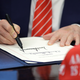 A close-up of Trump's hand and pen as he signs a document while wearing a striped red and white tie