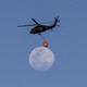 A firefighting helicopter carries a bucket as it flies past a nearly full moon.