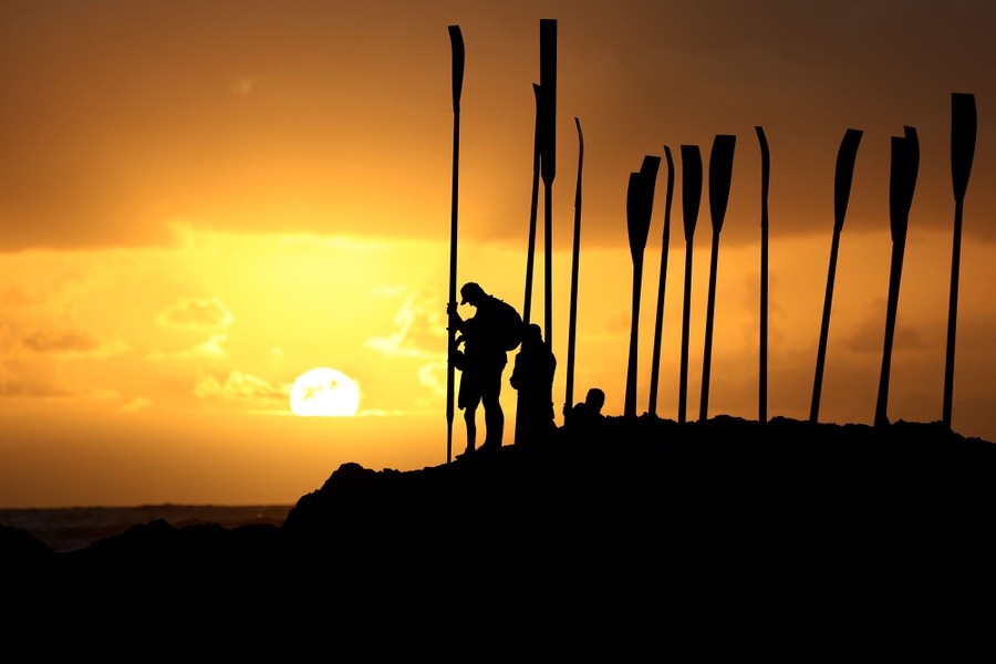 People hold long oars upright, silhouetted by the rising sun.