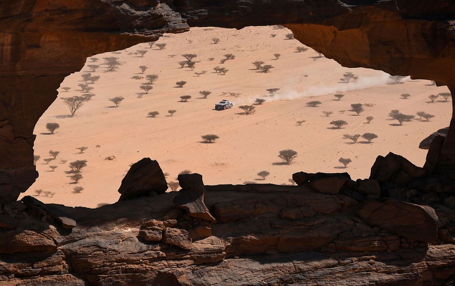 A rally car races through a desert, seen through a natural rock arch.