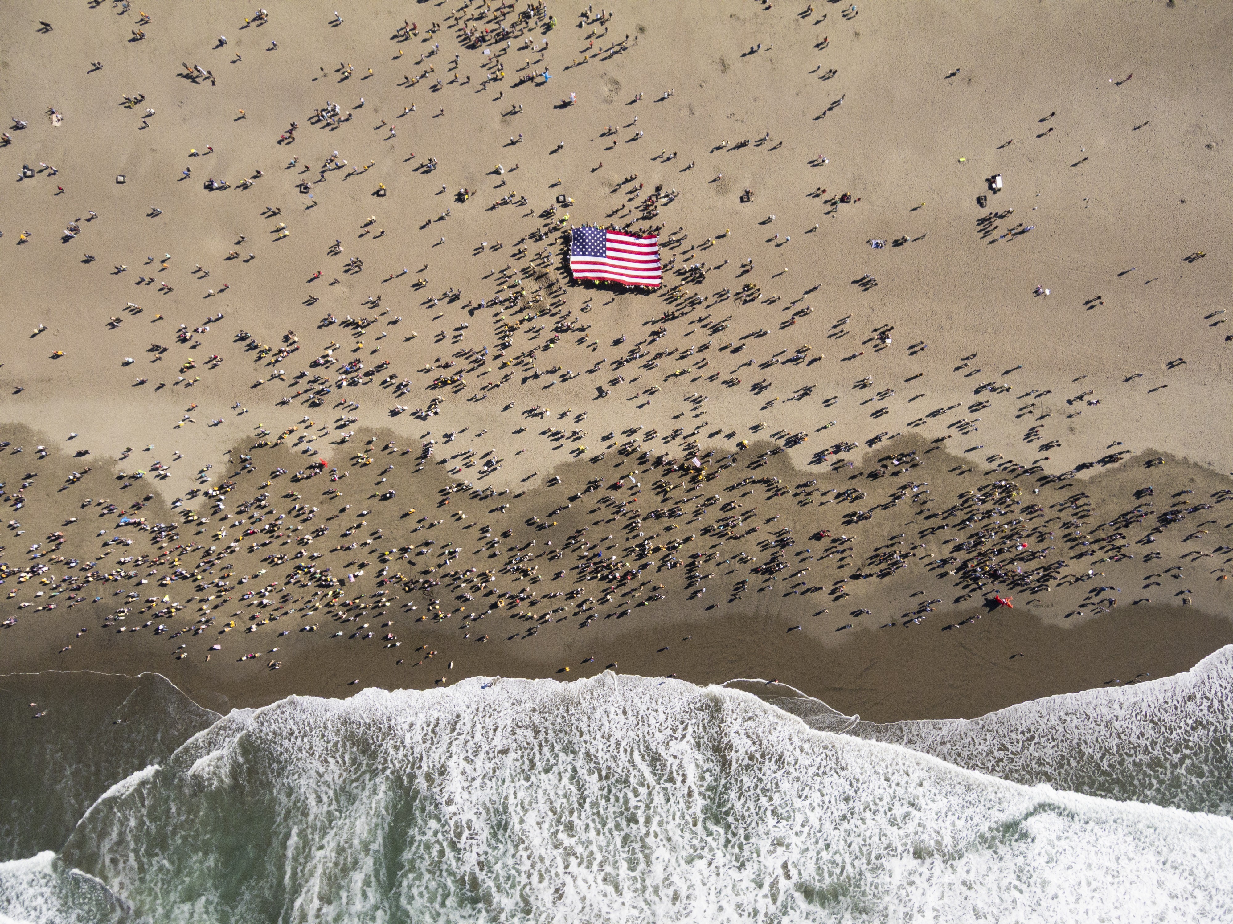 An aerial view of hundreds of people gathered on a beach, many holding up a large American flag.