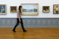 A woman, who is blurry, walks past a series of paintings in a museum.