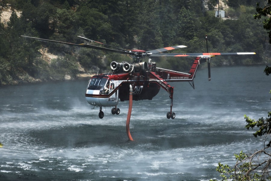 A helicopter hovers above a river, using a large hose to pump water aboard.