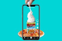 A phone records a hand spraying whipped cream into a can of Spaghetti-Os on top of a pizza.