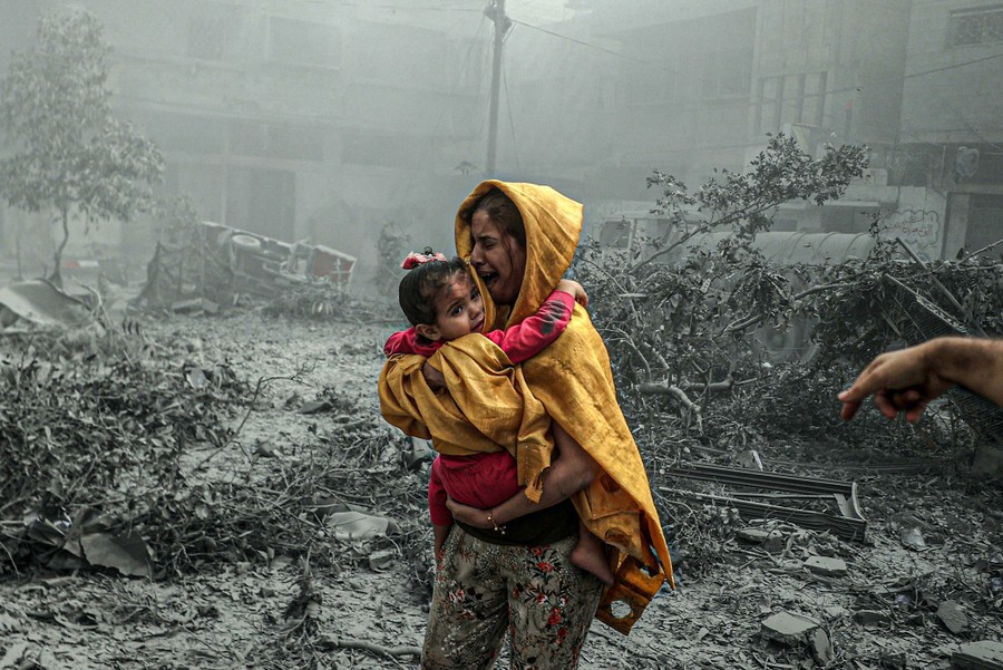 A woman carries a young girl through an open space in a city that is filled with bomb debris and covered in gray dust.