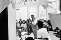 A black-and-white photo of a teacher teaching a class, captured through the window in the classroom door