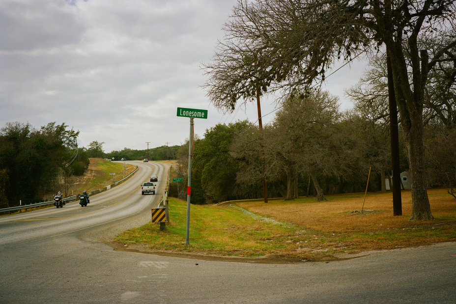 photo of intersection with green street sign for "Lonesome" street, with motorcycles and trucks driving past.