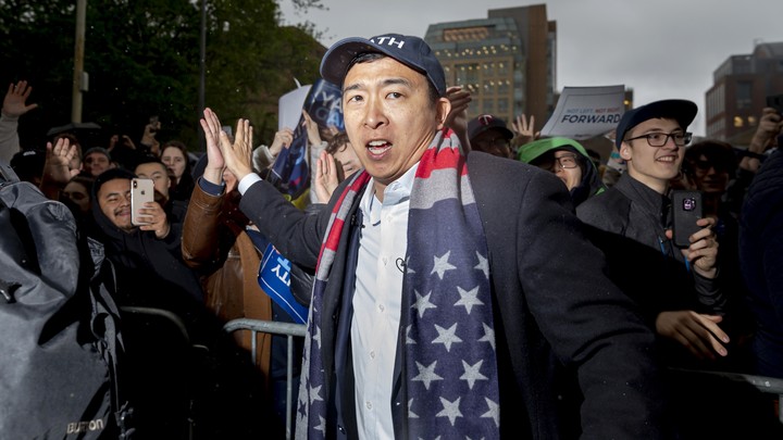 Andrew Yang high fives a supporter at a rally. An American flag is draped over his neck.