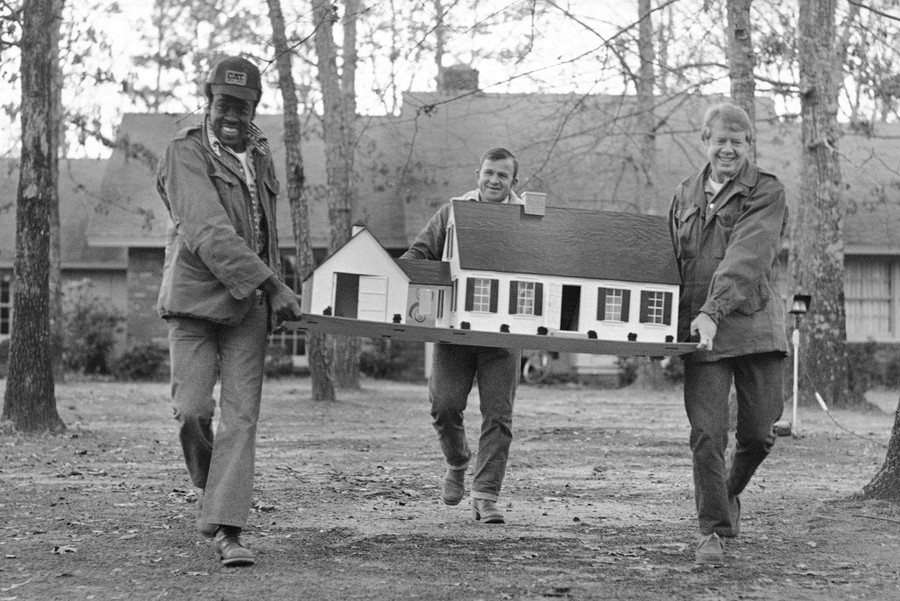 Three men carry a large dollhouse across a front lawn.