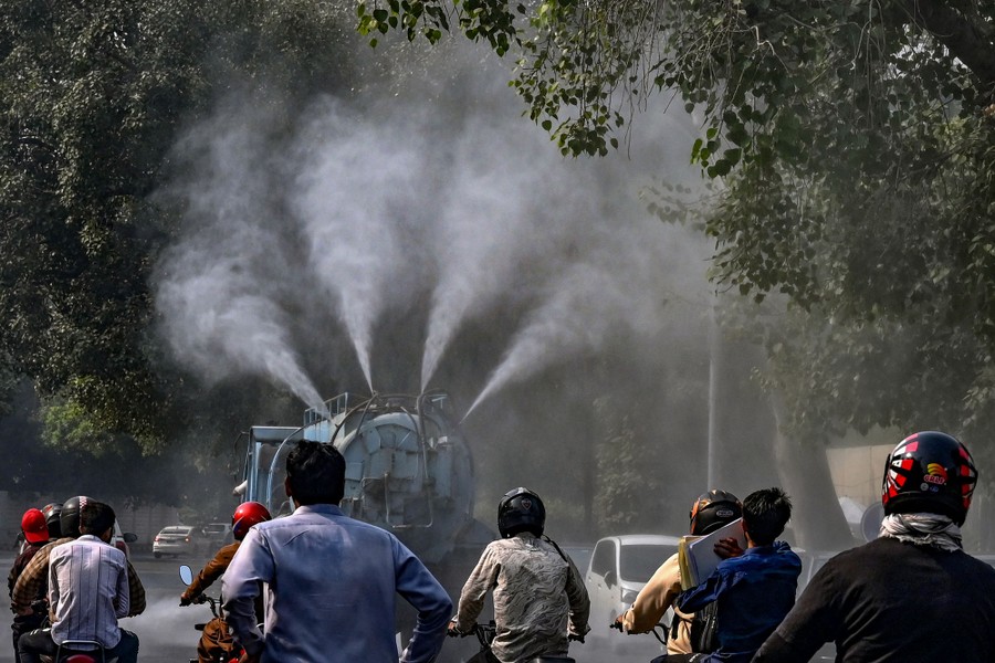 People on motorcycles follow behind a tanker truck that is spraying water up into the air on a road.