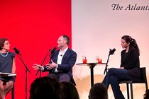 Three people sit onstage in black chairs with microphone stands beside each of them. The woman on the left is wearing a blue dress and blue necklace; she is looking at a man in a suit to her right who is gesticulating while he speaks, and another woman on the far right looks at them both and listens.