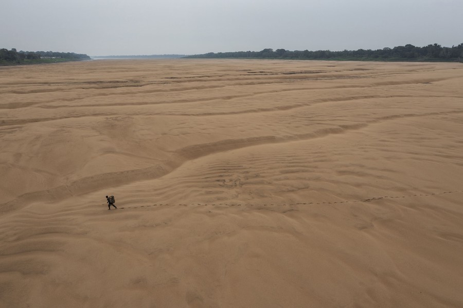 An elevated view of a person carrying a basket on their back while walking across a broad dry riverbed.