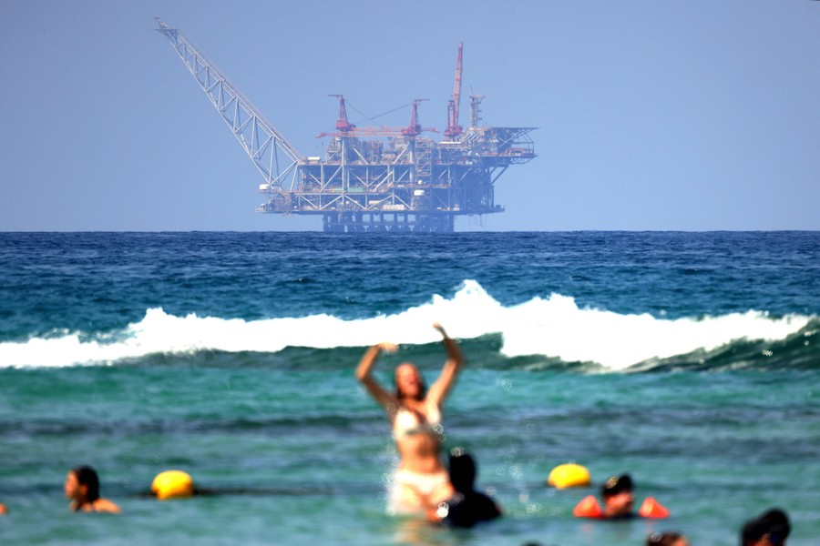 A large drilling platform in the background, with people playing in the surf at a beach in the foreground
