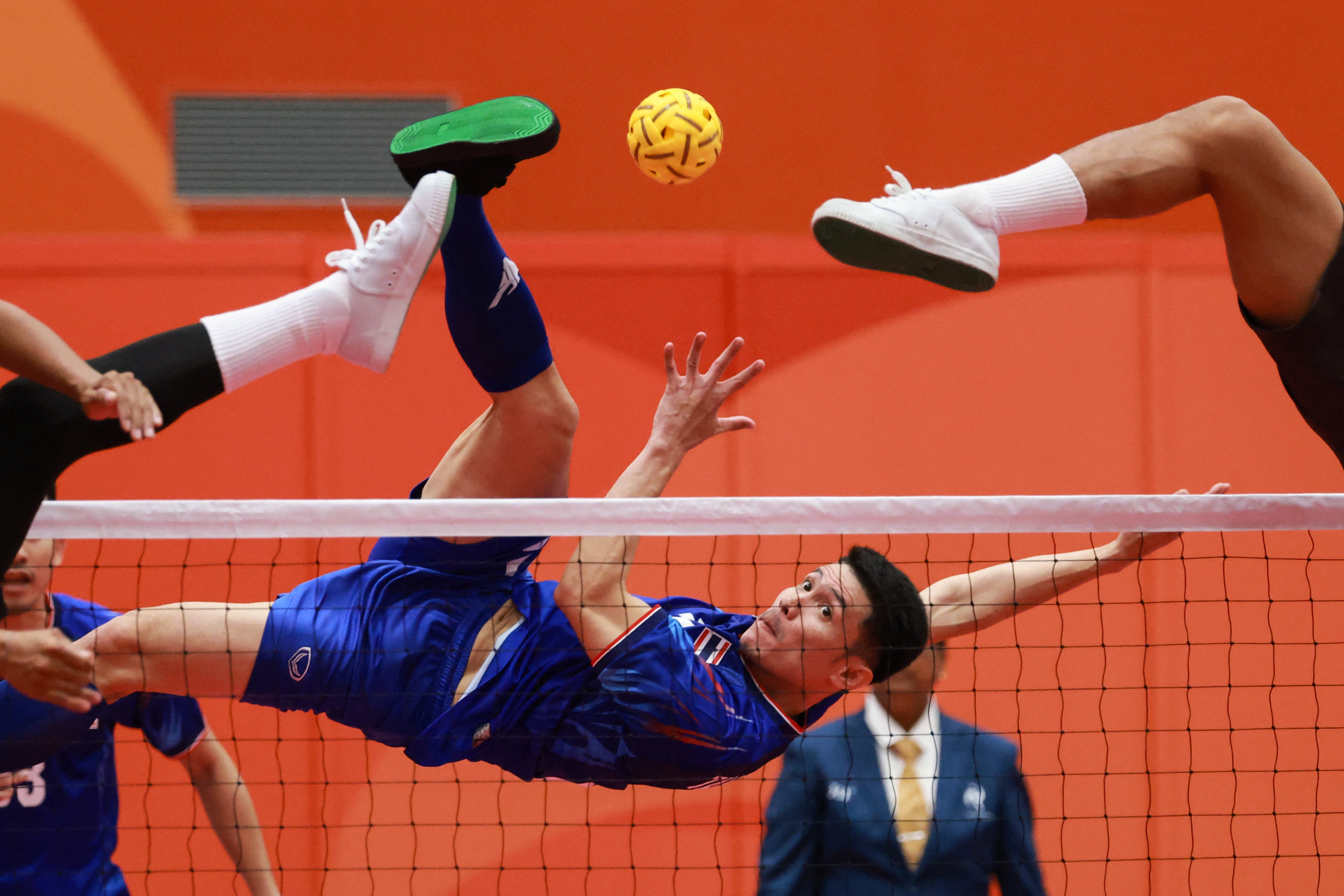 An athlete leaps high in the air to kick a small ball over a net, as defenders are seen with their feet also high in the air on the opposite side of the net.