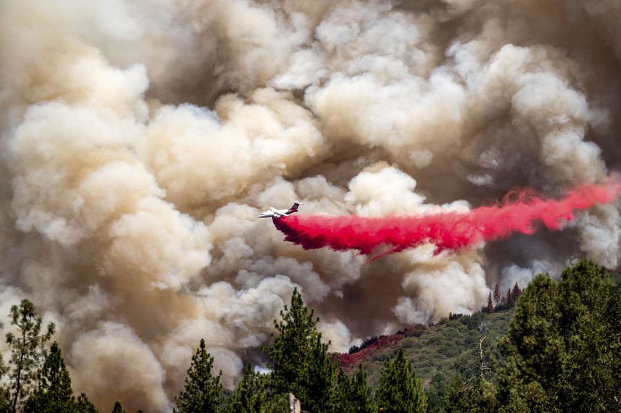 An air tanker drops red flame retardant while flying low in front of smoke rising from a burning forest.