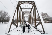 A man and a little boy hold hands while crossing a snowy bridge.