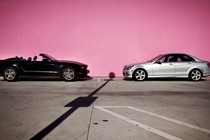 An image of a black convertible (left) and a silver sedan (right) that are parallel-parked along a pink wall, facing each other