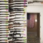 A photograph taken inside a gun shop, in which columns of rifles hang on white pegboard walls. An American flag is visible through an open doorawy.