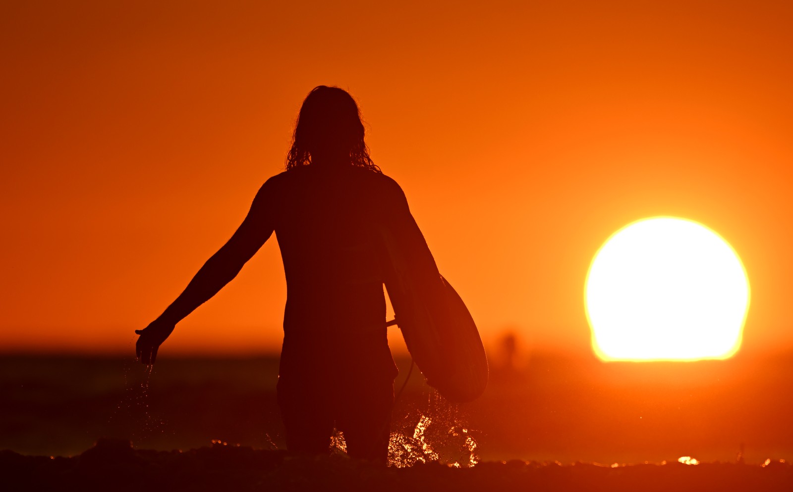 A person carrying a surfboard wades into the surf at sunset.