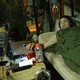 A man watches a movie on his laptop while wearing headphones and lying on a bed with camouflage bedding.