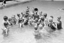 Children listen to a battery-powered radio receiver that is set on a table in a swimming pool.