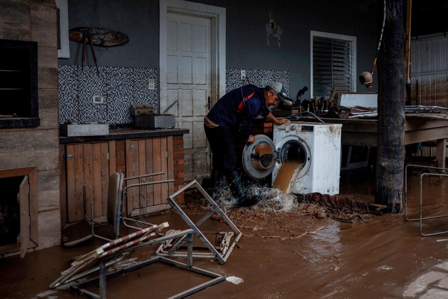 A person stands in a flood-damaged house, opening a washing machine that had been filled with muddy floodwater, which pours out.