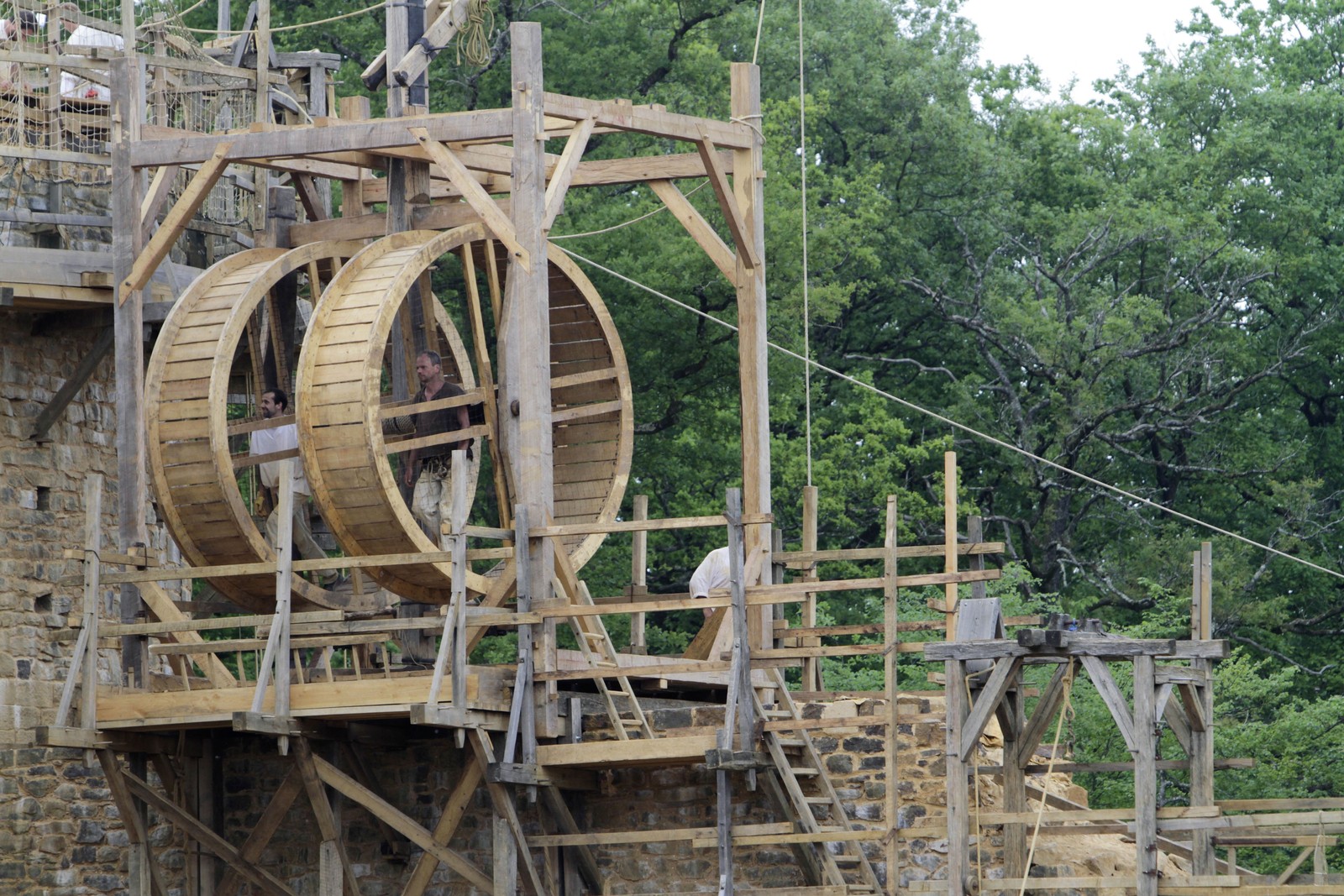 Two people walk inside the wheels of a medieval treadwheel crane, lifting material at a castle construction site.