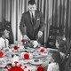 A black-and-white photo of a family sitting down for a holiday dinner, overlaid with red coronavirus particles