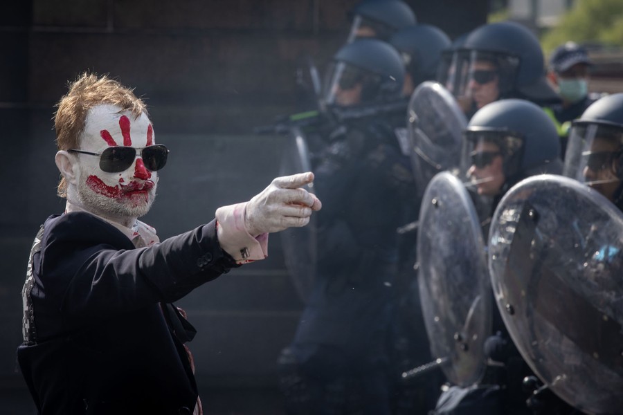 A protester with red and white face makeup points at riot police officers.