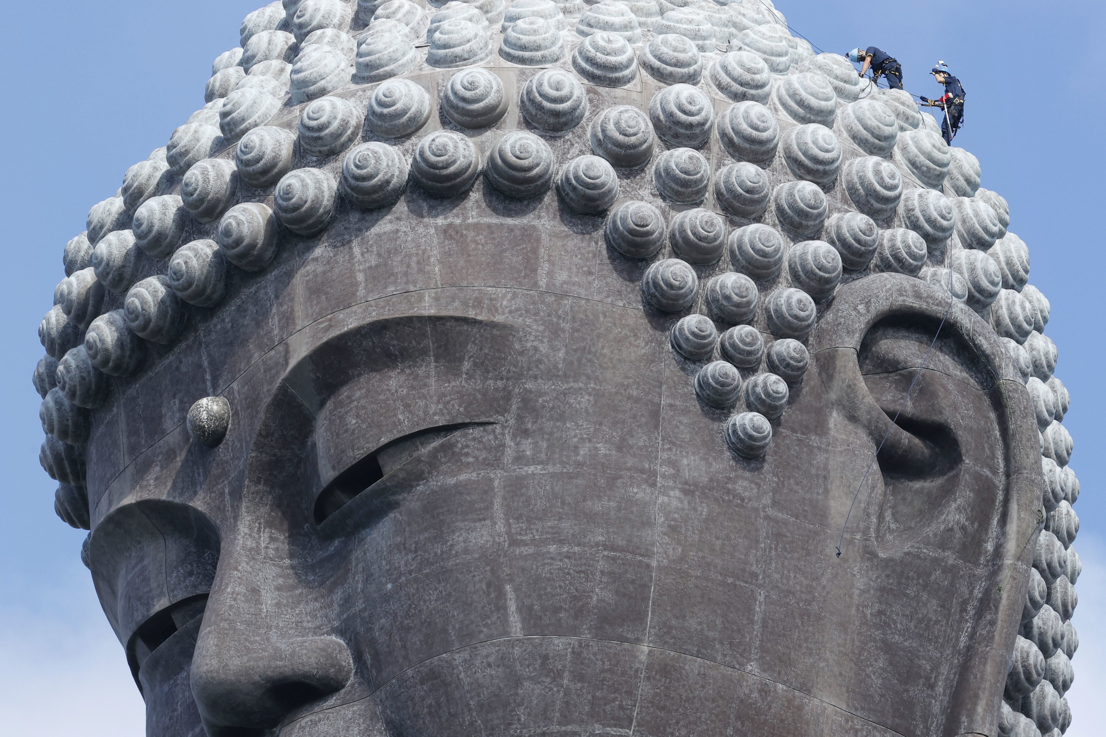 Two cleaners work on top of the head of a giant Buddha statue.